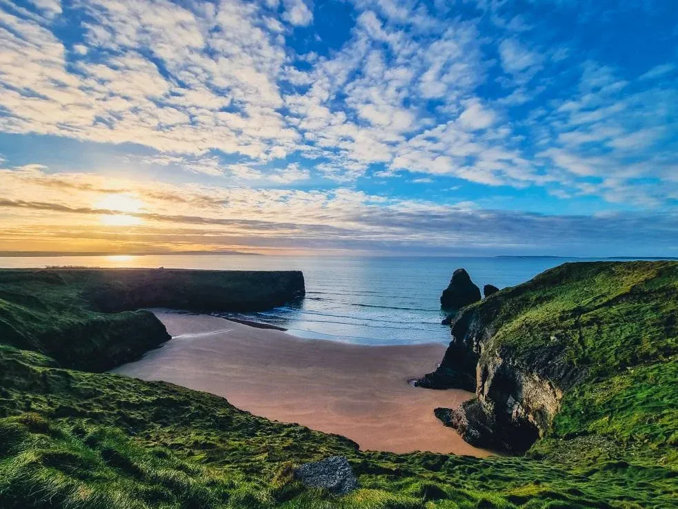 Scenic view of Ballybunion Beach in Ireland at sunset, with golden light reflecting on the sandy cove, rugged cliffs covered in green grass, and a partly cloudy sky over the calm Atlantic Ocean.