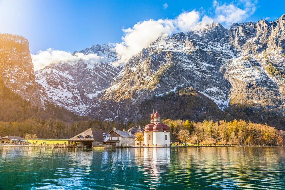 Serene alpine lake reflecting the snow-dusted peaks and red-domed church of Berchtesgaden National Park, surrounded by dense trees and wooden huts.