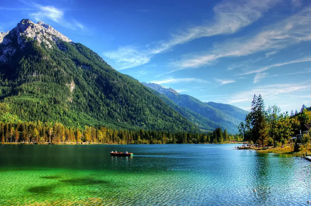 Scenic lake view in Berchtesgaden National Park with crystal-clear water, dramatic snow-capped mountains, and the red-domed Church of St. Bartholomew on the shore.