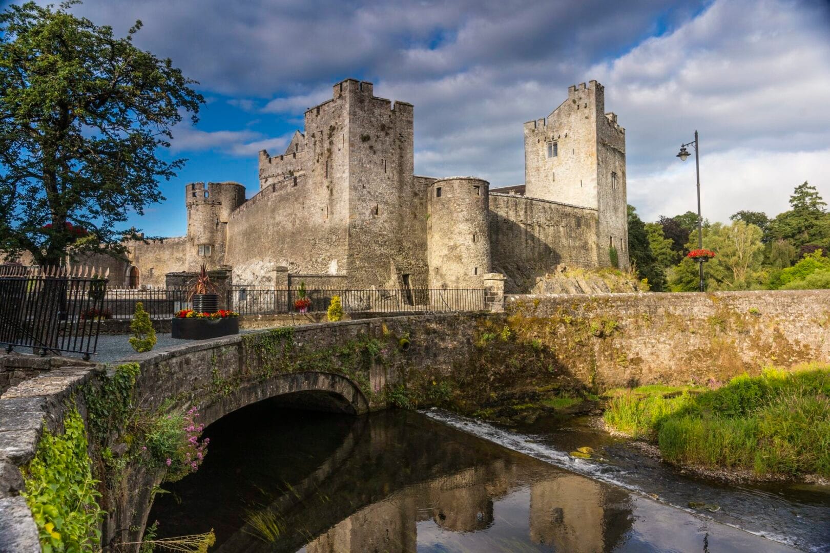View of Cahir Castle in Ireland, featuring medieval stone walls and towers beside a calm river with a stone bridge, surrounded by greenery and flowers under a partly cloudy sky.