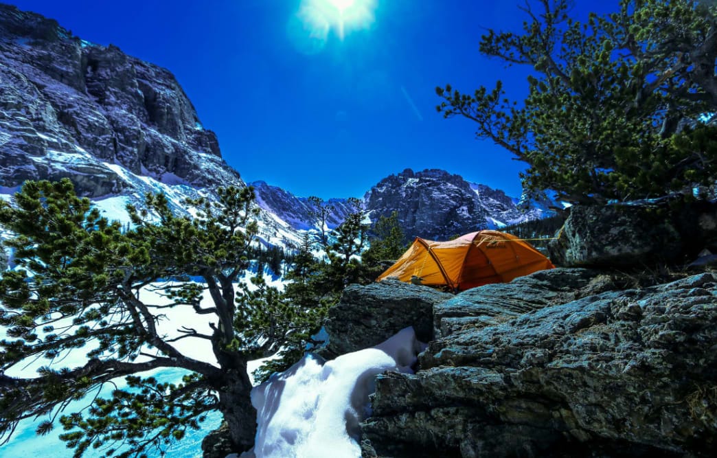 Bright orange tent set up on a snowy ledge with mountain views in Rocky Mountain National Park, Colorado, under a clear blue sky.