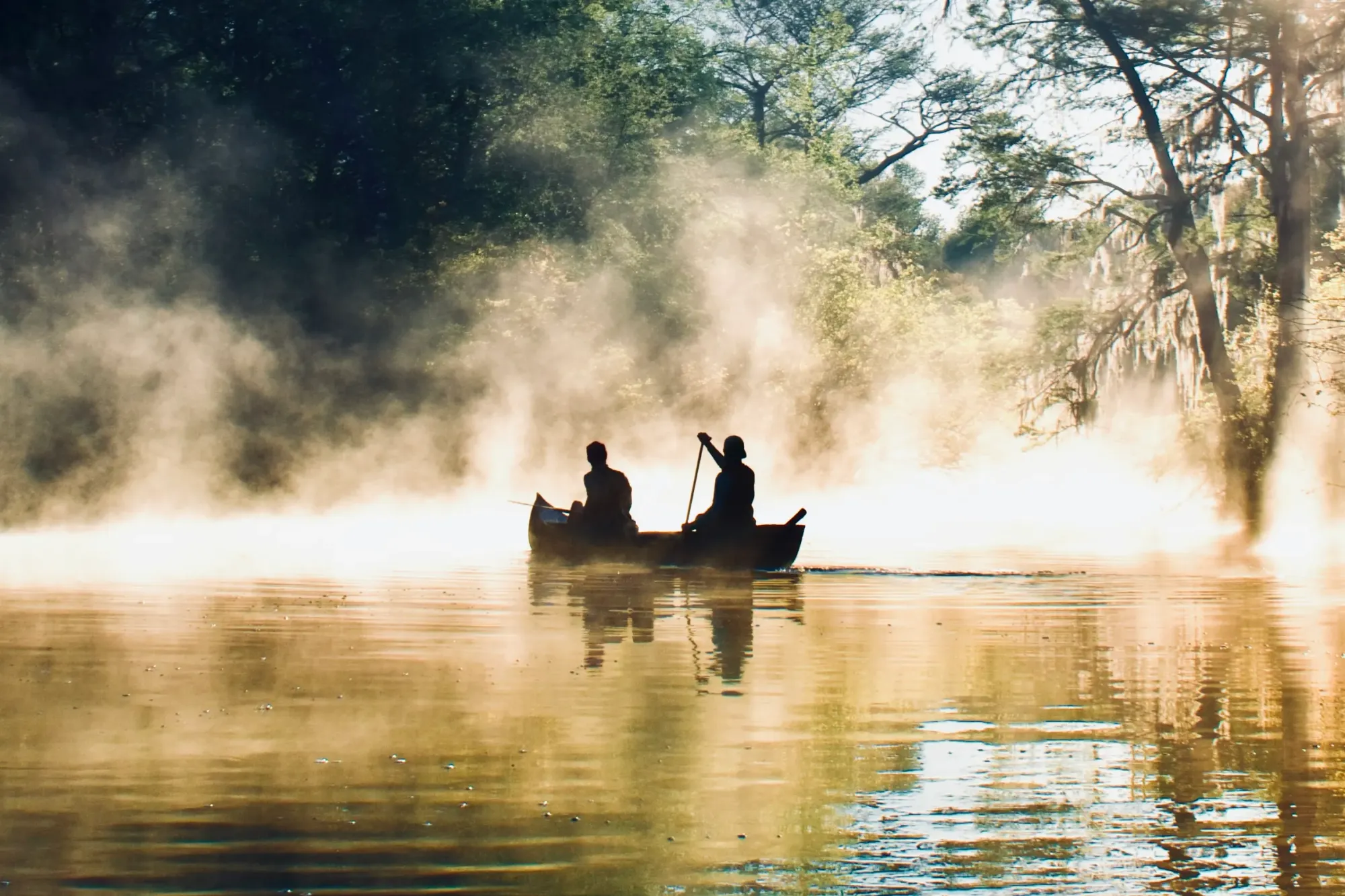 Two people canoe through a misty, golden-lit section of the Okefenokee Swamp surrounded by cypress trees and Spanish moss.