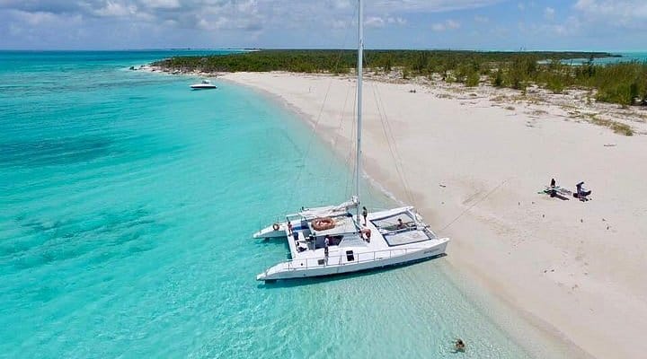 Catamaran anchored in the shallow, crystal-clear turquoise waters of Sapodilla Bay Beach in Turks and Caicos, with a white sandy shoreline, sparse greenery, and a few people relaxing on the beach under a partly cloudy sky