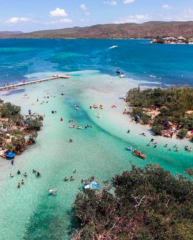 Aerial view of Cayo Aurora, also known as Gilligan’s Island, near Ponce, Puerto Rico, showing people swimming and relaxing in shallow, clear turquoise waters surrounded by lush mangroves, with a wooden pier extending into the bay and distant hills under a partly cloudy sky.