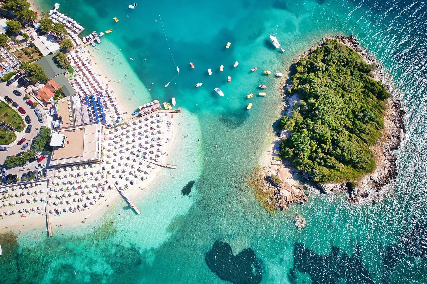 Aerial view of Ksamil beach and island in Albania with umbrellas, turquoise water, anchored boats, and lush greenery.