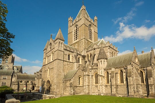 Exterior view of Christ Church Cathedral in Dublin, Ireland, showcasing its medieval stone architecture with pointed arches, detailed windows, and a central tower under a bright blue sky with scattered clouds.