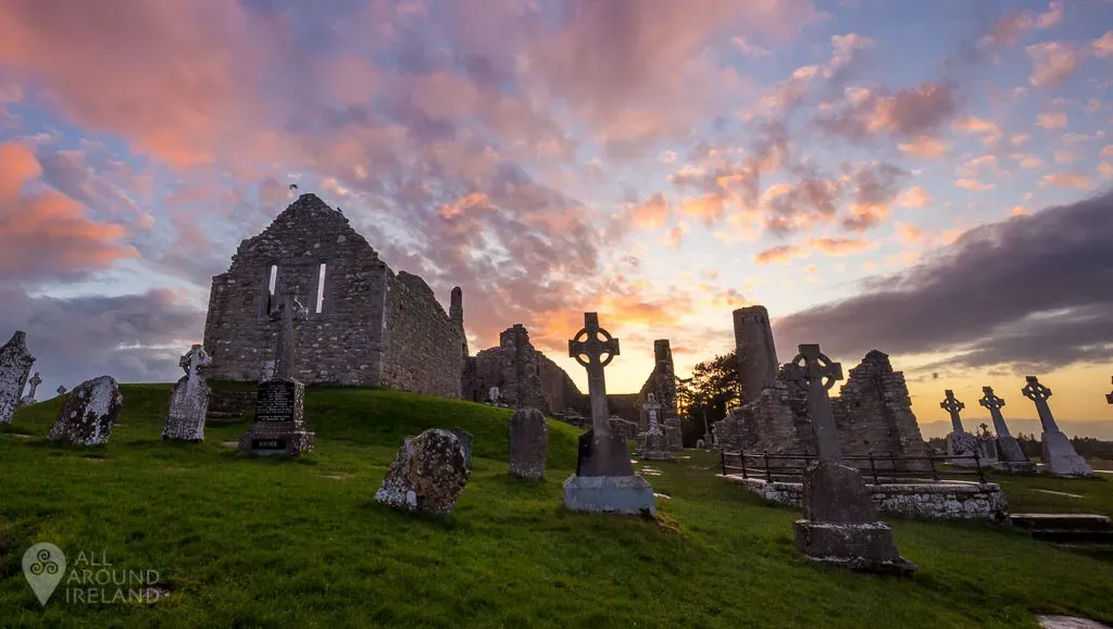 Ruins of the ancient monastic site of Clonmacnoise in County Offaly, Ireland, with Celtic crosses and old stone churches silhouetted against a colorful sunset sky.