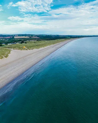 Aerial view of Curracloe Beach in Wexford, Ireland, featuring a long stretch of sandy shoreline bordered by calm blue ocean waters and grassy dunes under a partly cloudy sky.