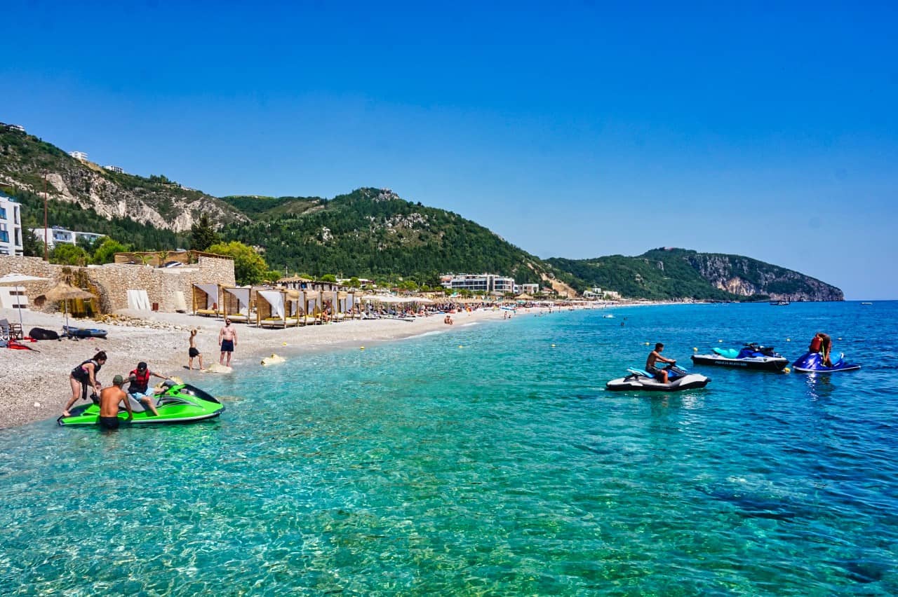 Dhërmi Beach in Albania with crystal-clear water, jet skis, sunbeds, beachgoers, and a scenic mountain backdrop.