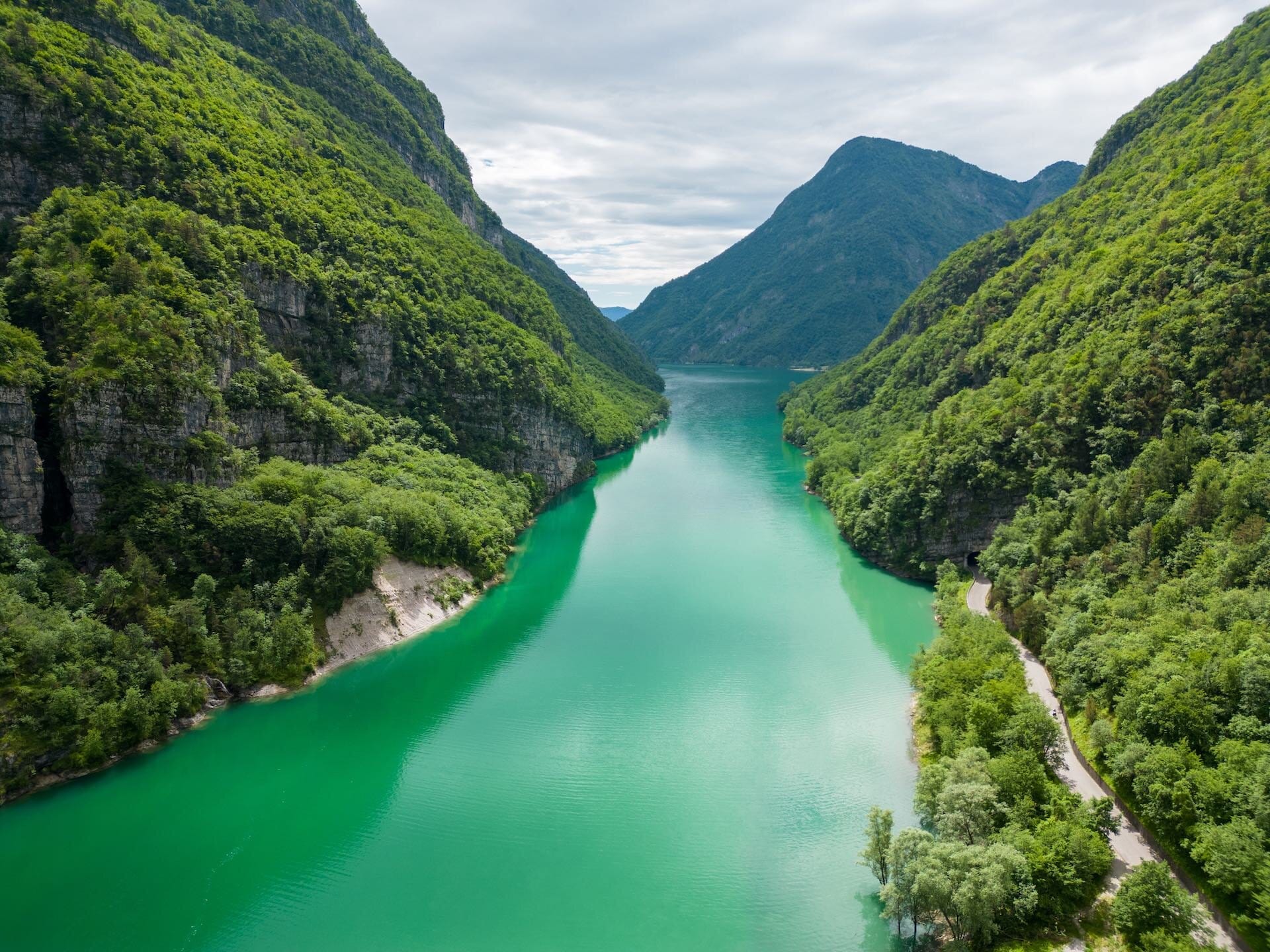 A winding river flanked by steep forested mountains in Dolomiti Bellunesi National Park, Italy, with a cloudy sky overhead and a scenic road along the water’s edge.