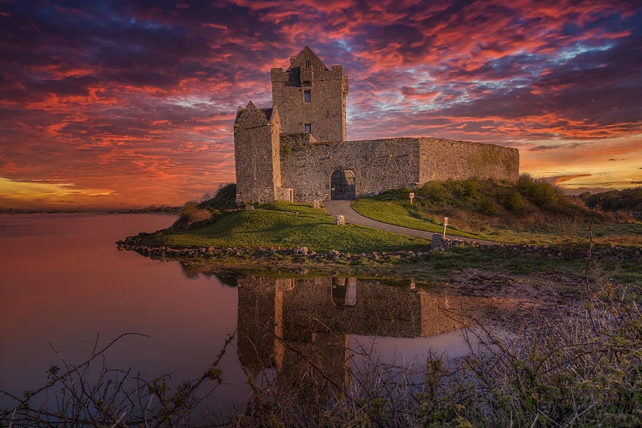Dunguaire Castle in Ireland at sunset, with dramatic purple and orange clouds reflected in the calm water surrounding the stone fortress on a grassy shoreline.