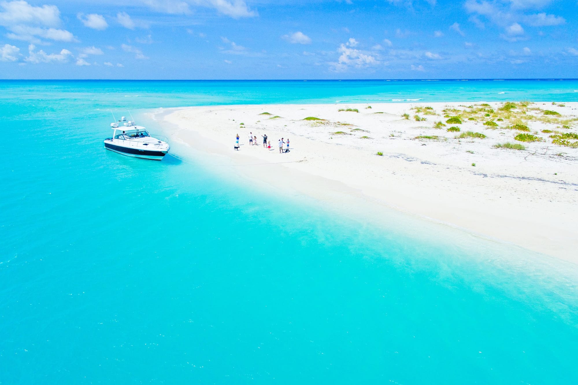 A yacht anchored near the white sandy shore of Fort George Cay in Turks and Caicos, with a small group of people and a dog standing on the beach, surrounded by clear turquoise waters under a sunny blue sky.