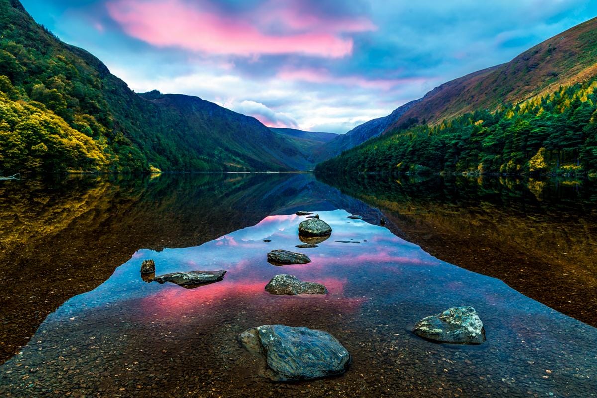 Scenic view of Glendalough Lake in Ireland, with calm waters, colorful sunset skies, surrounding green hills, and rocks in the foreground."