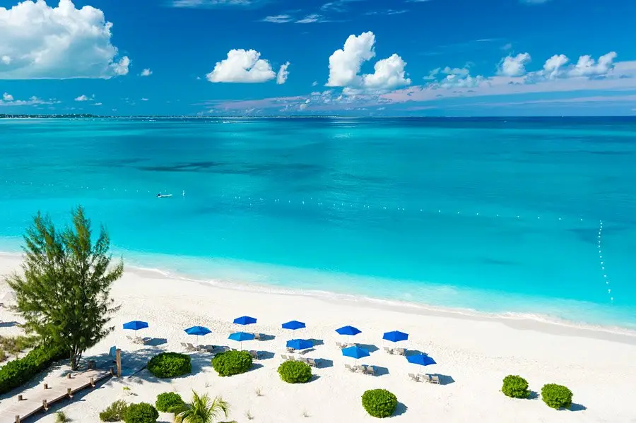 Scenic view of Grace Bay Beach in Turks and Caicos featuring soft white sand, rows of blue beach umbrellas and lounge chairs, turquoise ocean water, and a few scattered clouds in a bright blue sky.