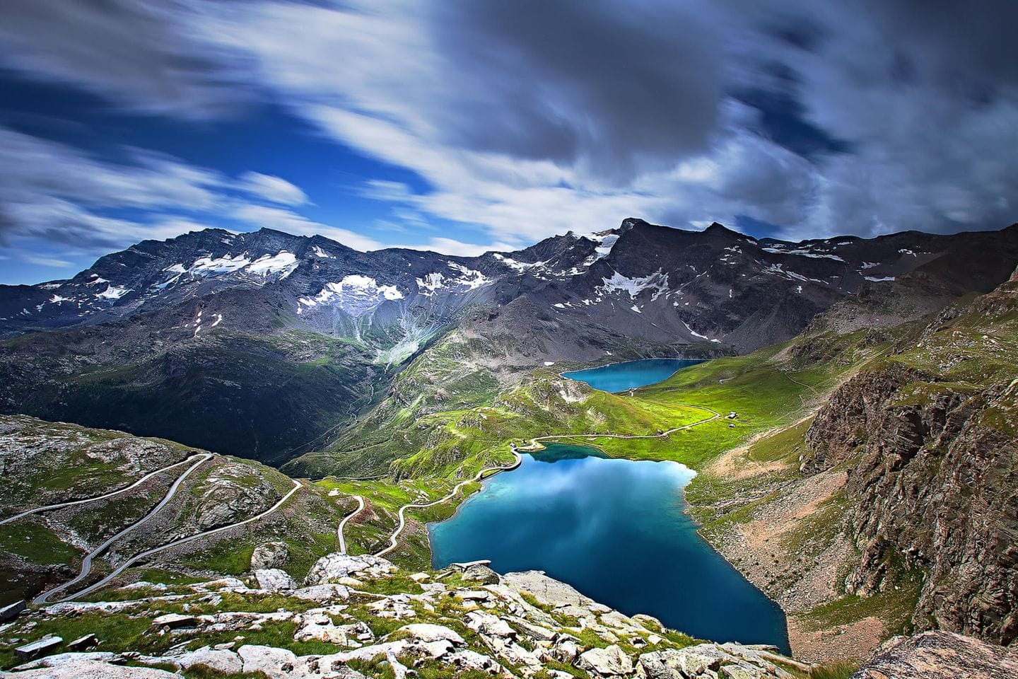 Scenic landscape of Gran Paradiso National Park featuring emerald alpine lakes, green valleys, and snow-capped mountains under a dramatic cloudy sky.