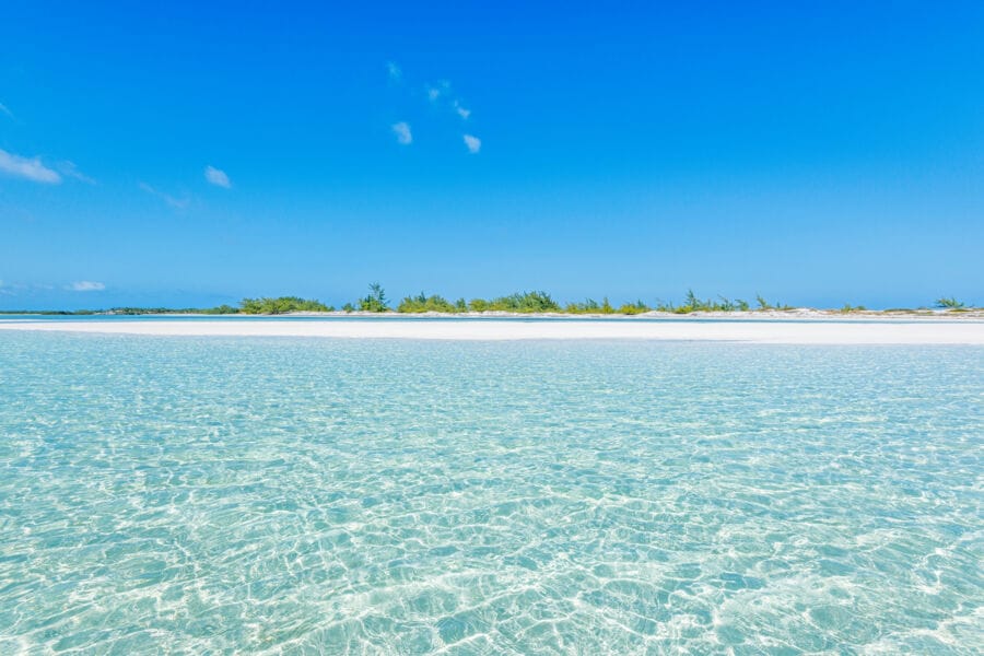 Crystal-clear shallow waters leading to a white sand beach with sparse greenery at Half Moon Bay in Turks and Caicos, under a vibrant blue sky with minimal clouds.