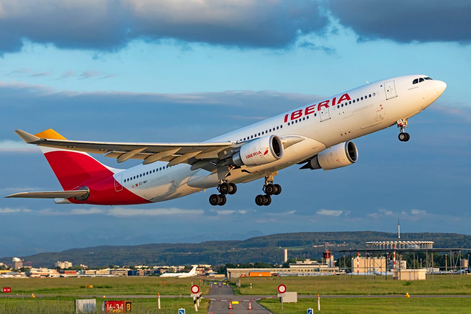 Iberia Airbus A330 taking off from runway with cityscape and hills in the background.