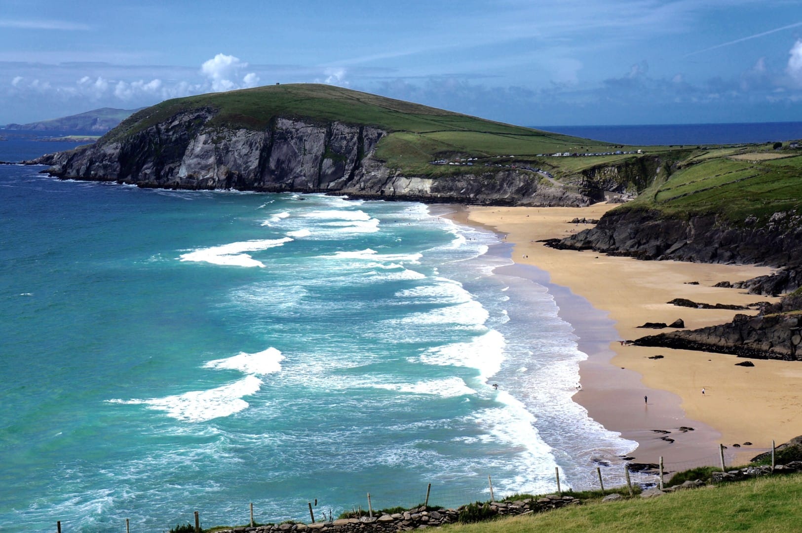 Scenic view of Inch Beach in Ireland, featuring turquoise ocean waves rolling onto a wide sandy shore, surrounded by rugged cliffs and lush green hills under a partly cloudy blue sky