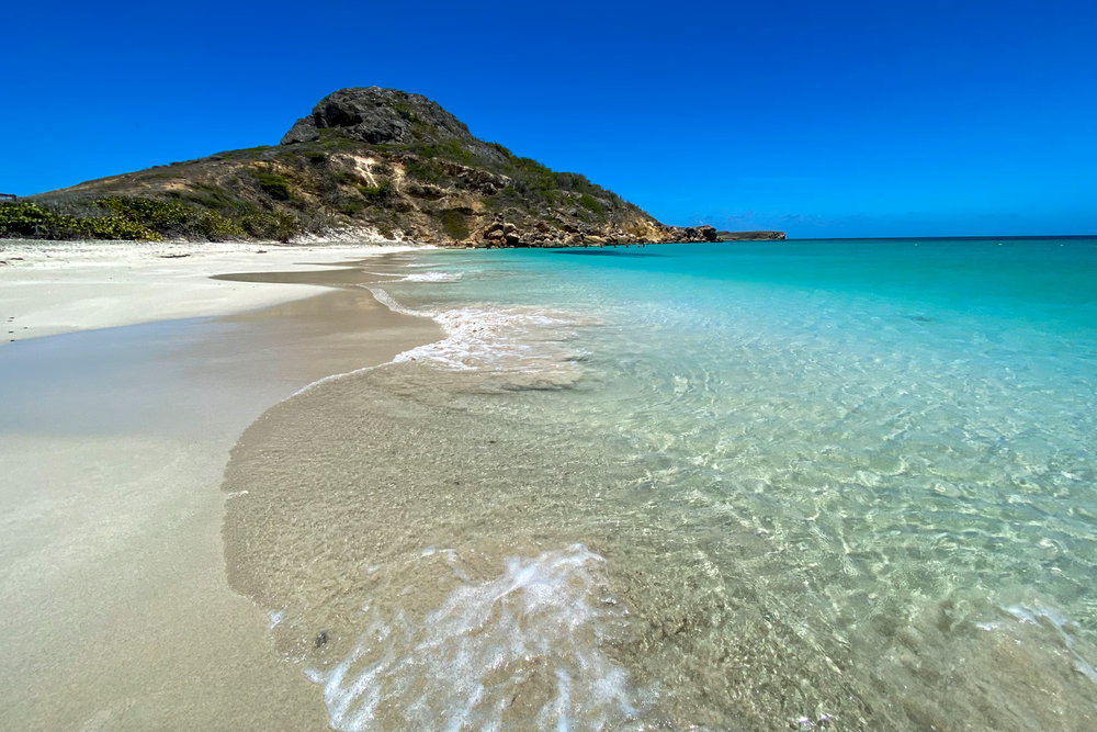 Scenic beach at Isla Caja de Muertos featuring crystal-clear shallow waters, a pristine sandy shoreline, and a rugged green hill under a bright blue tropical sky.