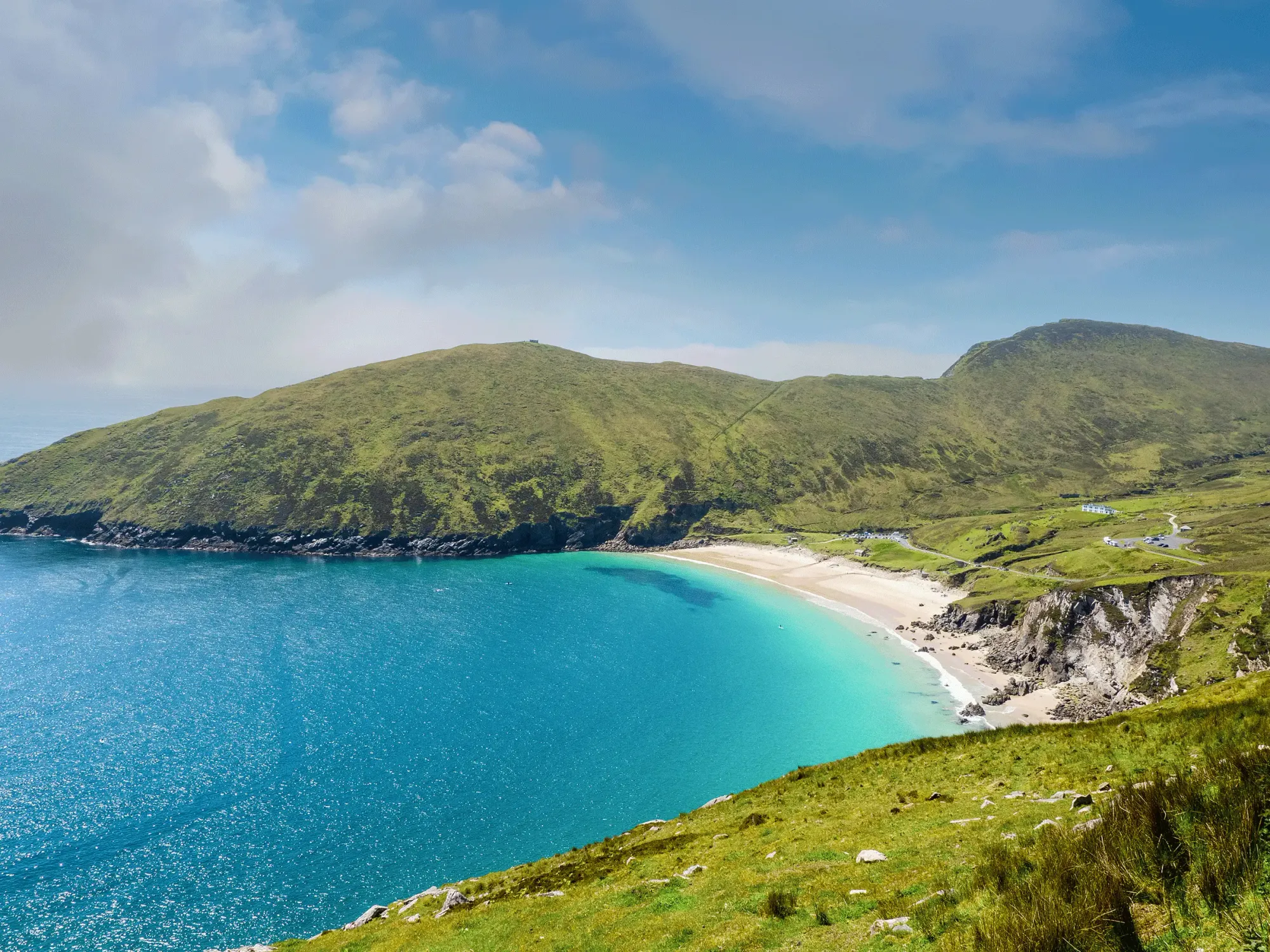 Stunning aerial view of Keem Bay Beach on Achill Island, Ireland, featuring a crescent-shaped sandy beach, vibrant turquoise waters, and rolling green hills under a bright blue sky with scattered clouds.