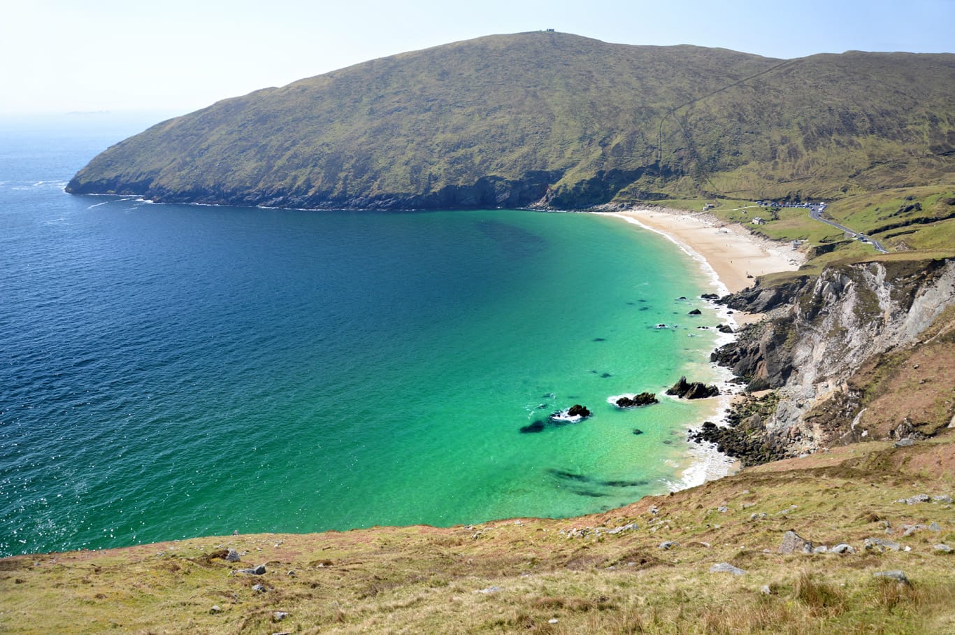 Scenic view of Keem Bay at Achill Island, featuring turquoise waters, sandy beach, rocky cliffs, and grassy hills under a clear sky.