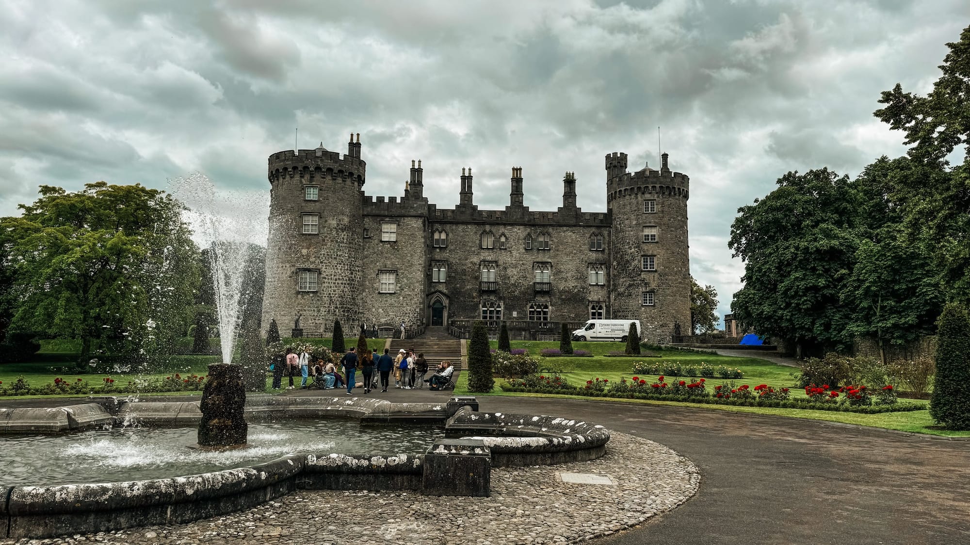Front view of Kilkenny Castle in Ireland with a large stone fountain in the foreground, surrounded by manicured gardens, red flowers, and groups of visitors under a cloudy sky.