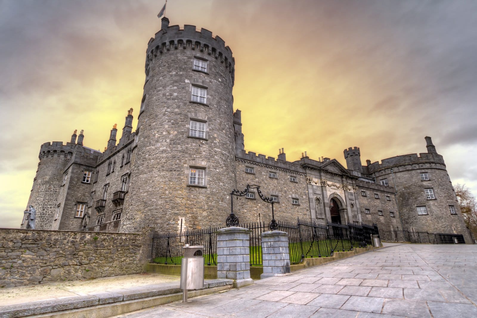 Front view of Kilkenny Castle in Kilkenny, Ireland, featuring its round stone towers and fortified walls under a dramatic, golden sunset sky.