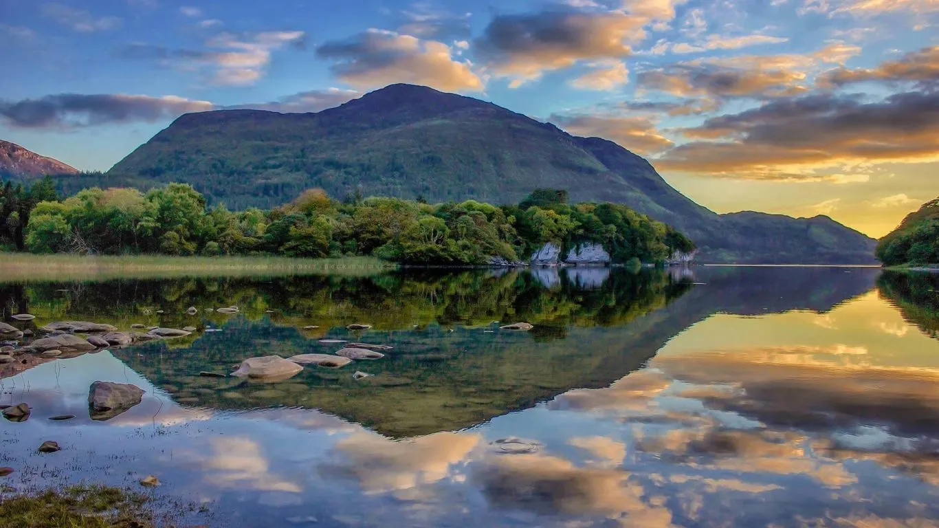 Peaceful sunset view at Killarney National Park, Ireland, showing calm lakes, tree-lined shores, rocky edges, and a mountain silhouette.