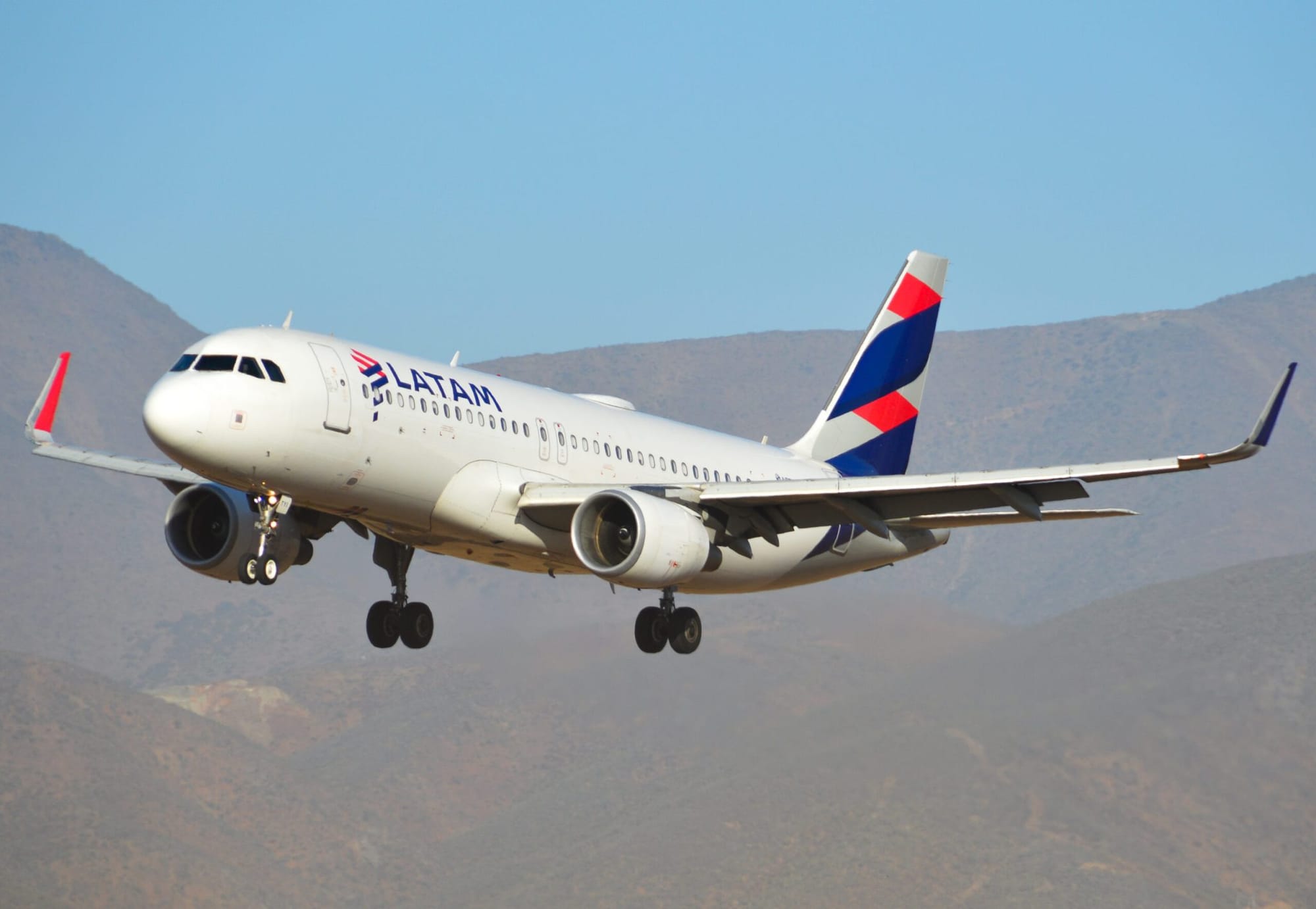 LATAM Airlines Airbus A320 descending with landing gear extended against a backdrop of dry, mountainous terrain under a clear blue sky.