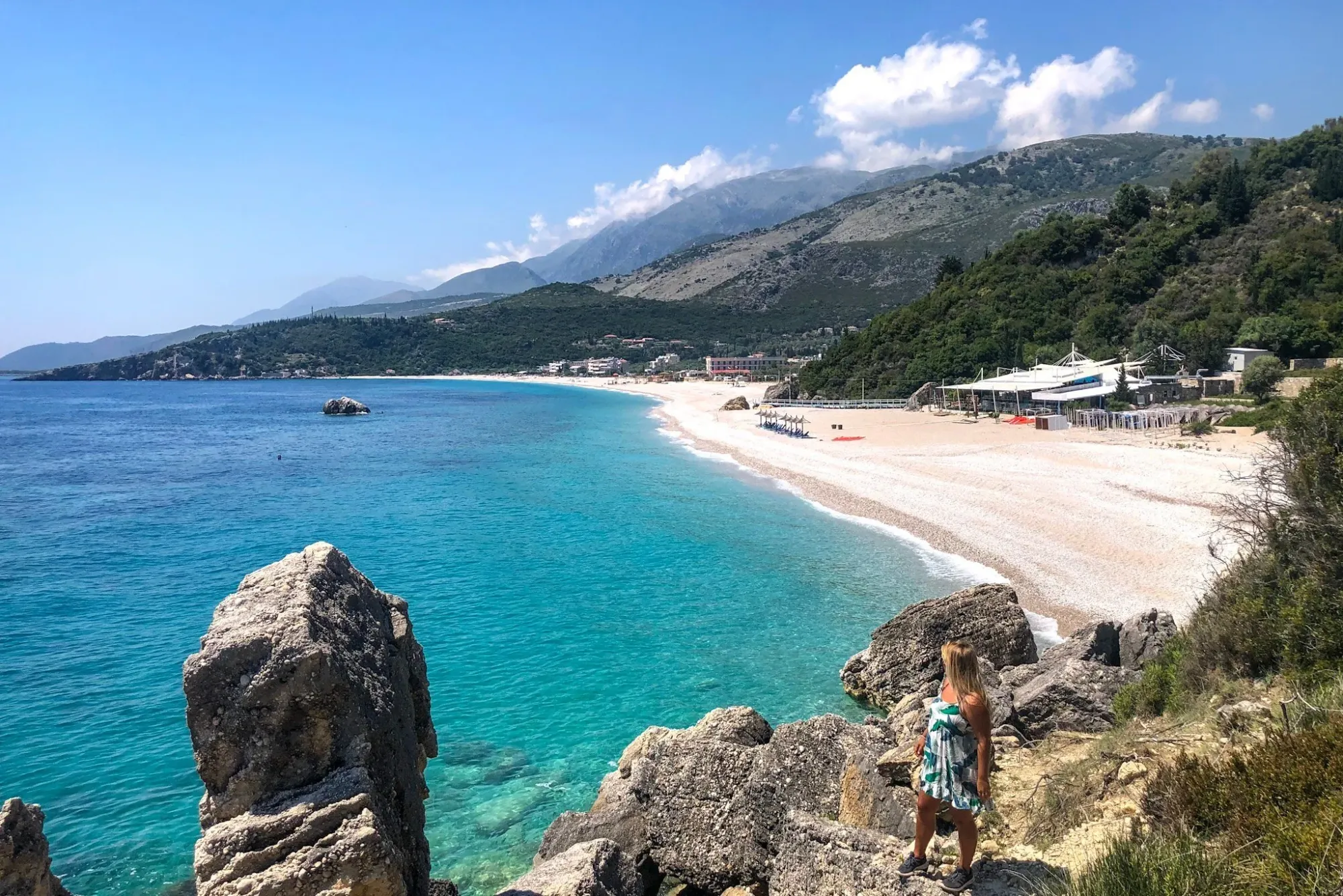 Livadhi Beach near Dhërmi, Albania with clear blue water, pebbled shore, mountain views, and a visitor standing on coastal rocks.