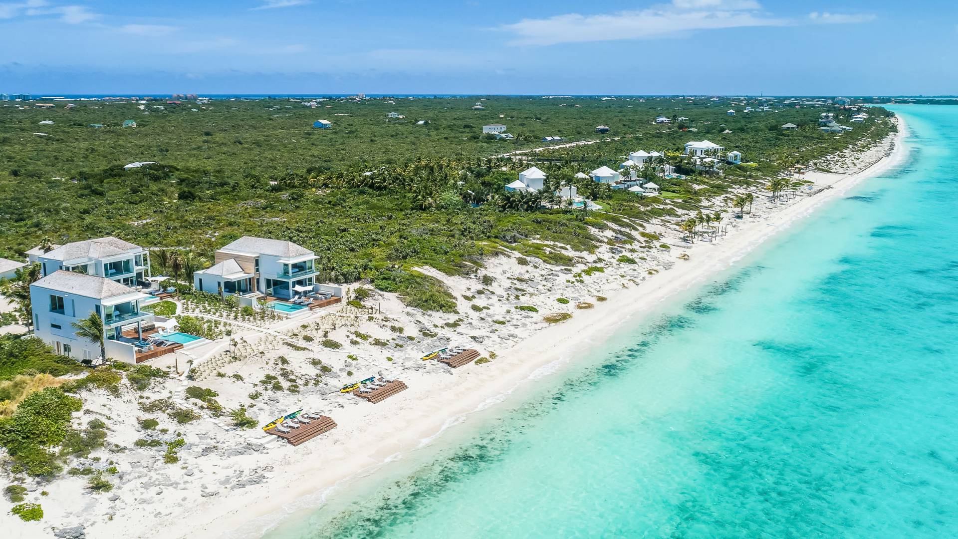 Aerial view of Long Bay Beach in Turks and Caicos featuring luxury beachfront villas with private pools, white sandy shoreline, turquoise ocean water, and lush green vegetation extending inland under a clear blue sky.