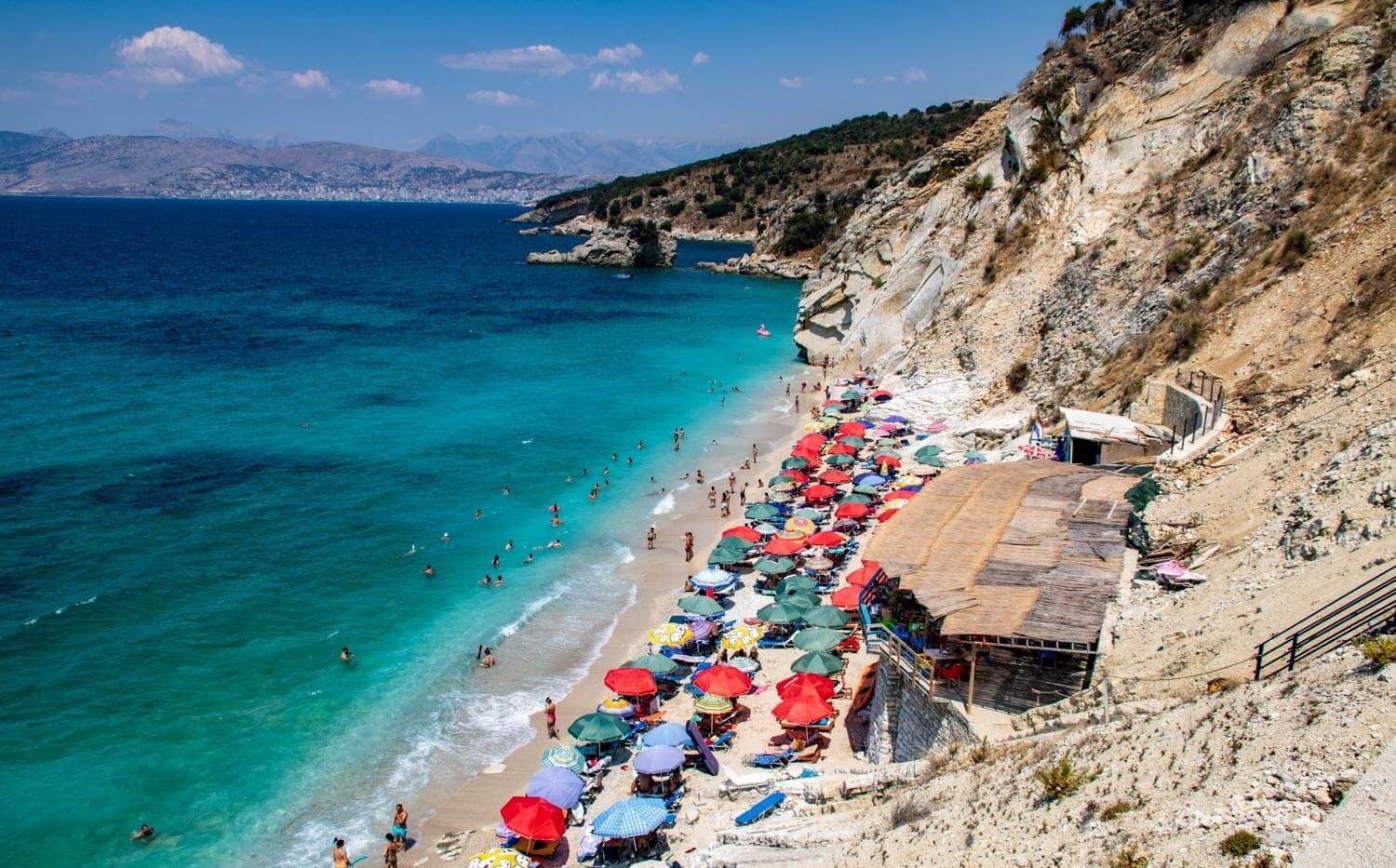 Manastir Beach in Albania with colorful umbrellas, turquoise water, white cliffs, and swimmers enjoying the clear shoreline.