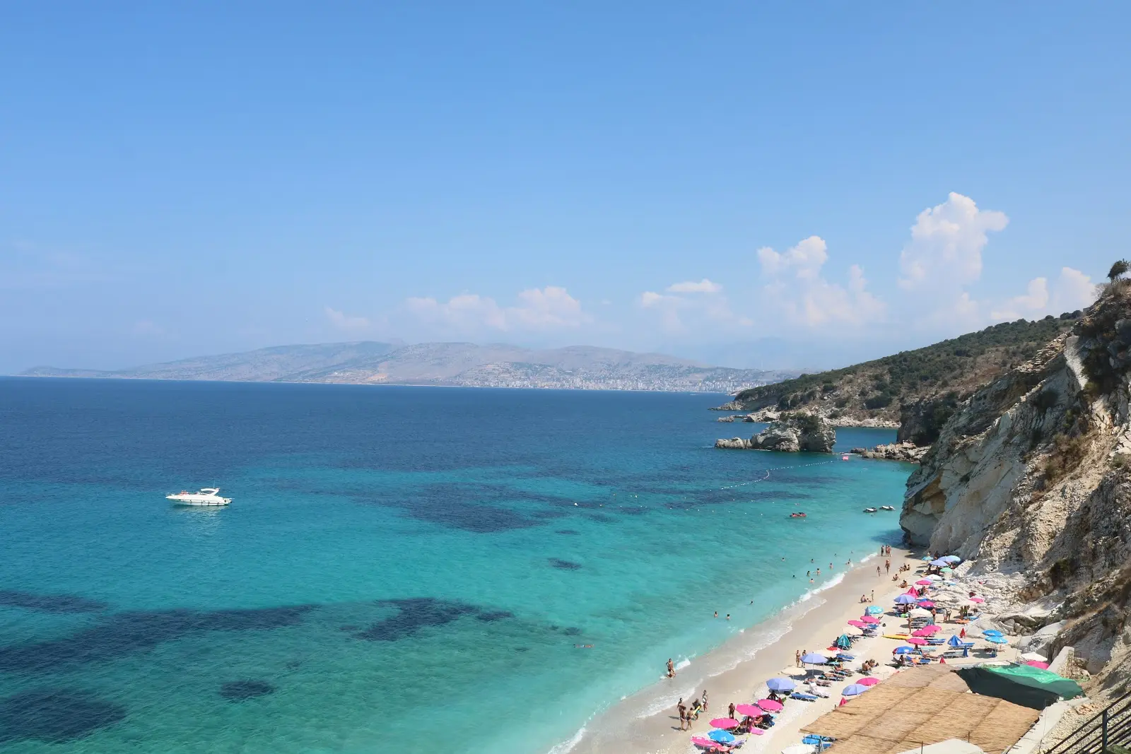 Mirror Beach in Albania with white cliffs, colorful umbrellas, turquoise water, swimmers, and a boat anchored near the shore.