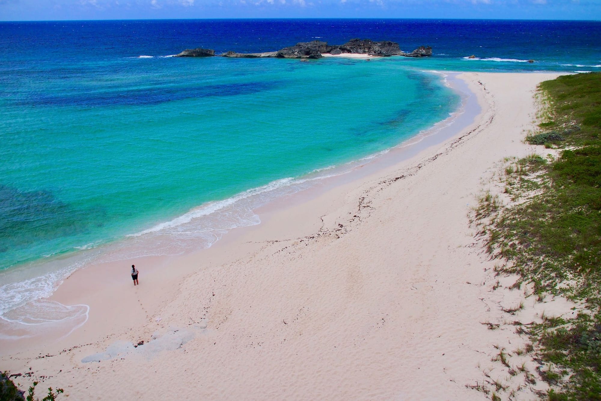 View of Mudjin Harbor Beach in Turks and Caicos showing a person standing on soft white sand near turquoise and deep blue ocean waters, with rocky outcroppings and light vegetation along the shoreline under a partly cloudy sky.