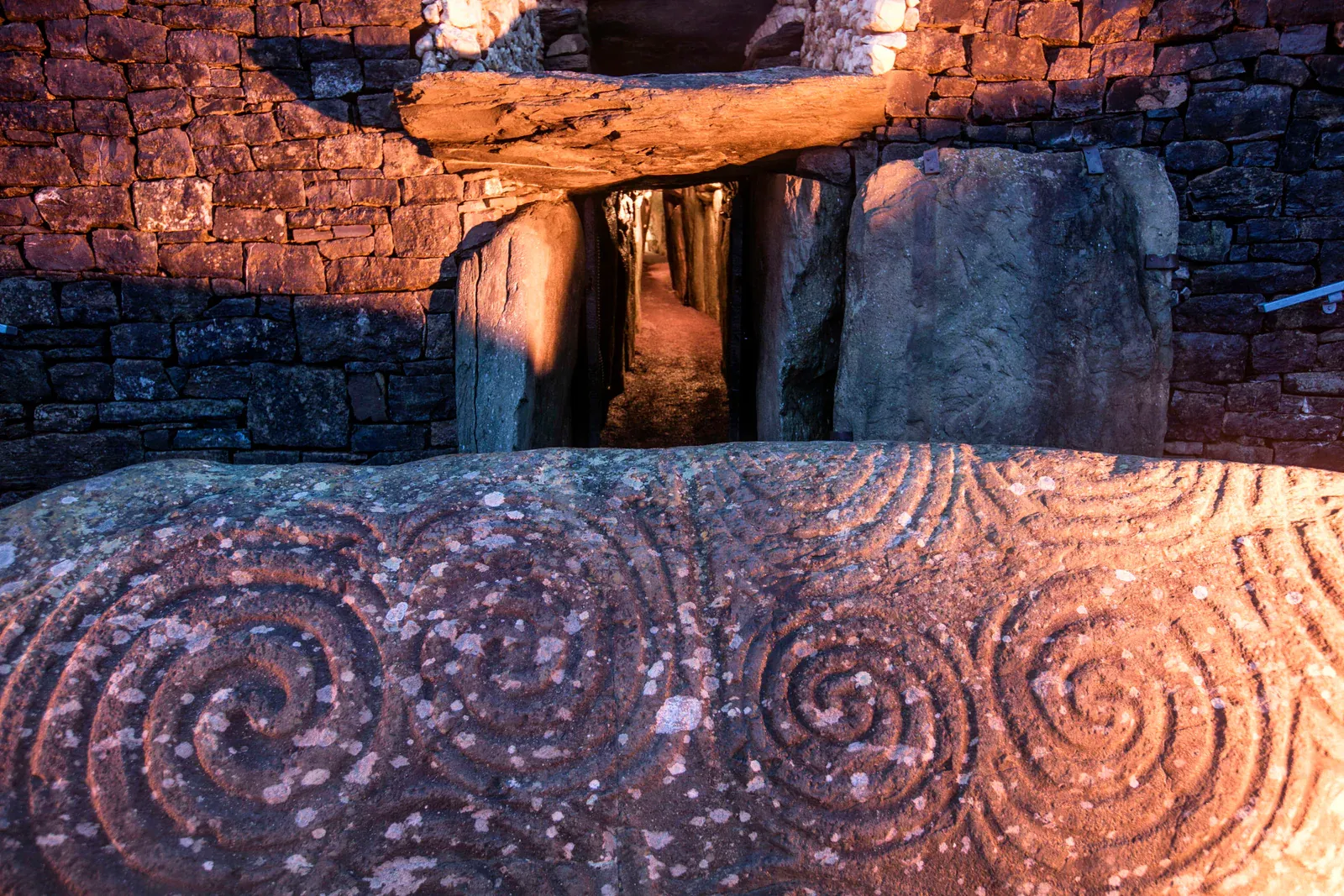 Entrance to Newgrange, a prehistoric passage tomb in County Meath, Ireland, featuring large stone slabs with intricate spiral carvings illuminated by warm light against a backdrop of ancient stone walls.
