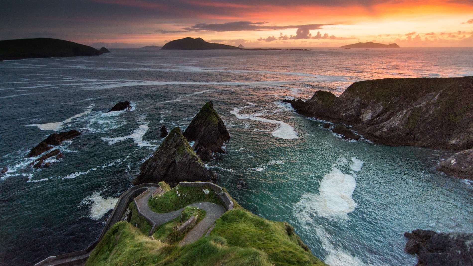 Scenic sunset view of rocky coastline and winding path at Dunquin Pier in Dingle, Ireland, with dramatic ocean waves and distant islands.