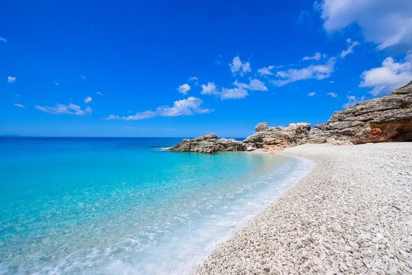 Palasa Beach in Albania with white pebbles, crystal-clear turquoise water, rocky shoreline, and a bright blue sky.