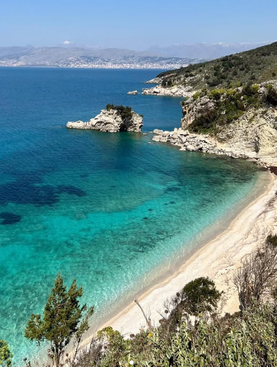 Mirror Beach (Pasqyra) in South Albania with clear turquoise water, rocky cliffs, and a narrow white-sand shoreline.