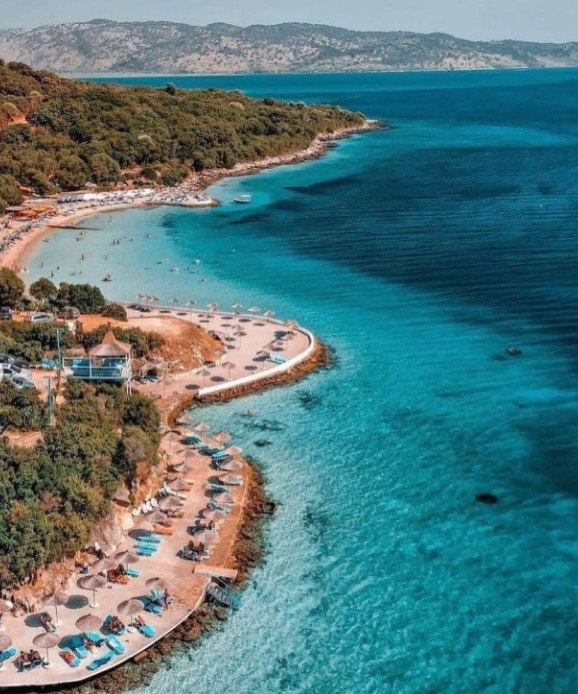 Pema e Thatë Beach in Ksamil, Albania, with sunbeds, umbrellas, lush greenery, and clear turquoise water along a curved coastline.