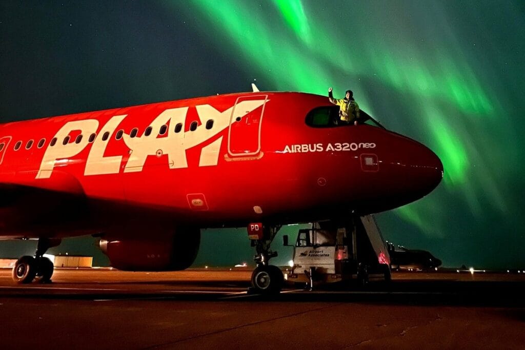 A solid red plane with Play on the side at night under the bright green northern lights in the sky in Iceland.