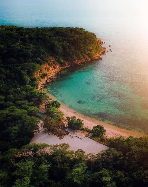 Aerial view of Playa Jaboncillo Beach near Ponce, Puerto Rico, showing a small secluded cove with clear turquoise waters, a narrow white sandy beach, and dense green forest covering the surrounding cliffs under soft sunlight.