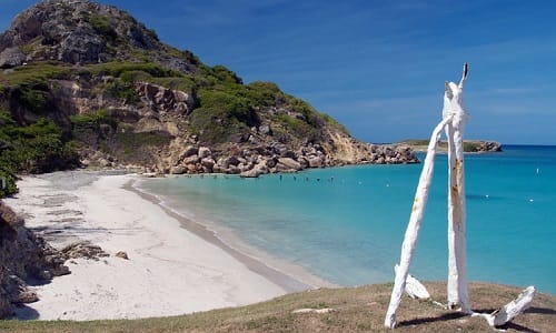 View of Playa Tamarindo featuring a white sandy beach, turquoise waters, rocky cliffs covered in greenery, and a rustic white wooden sculpture in the foreground under a clear blue sky.
