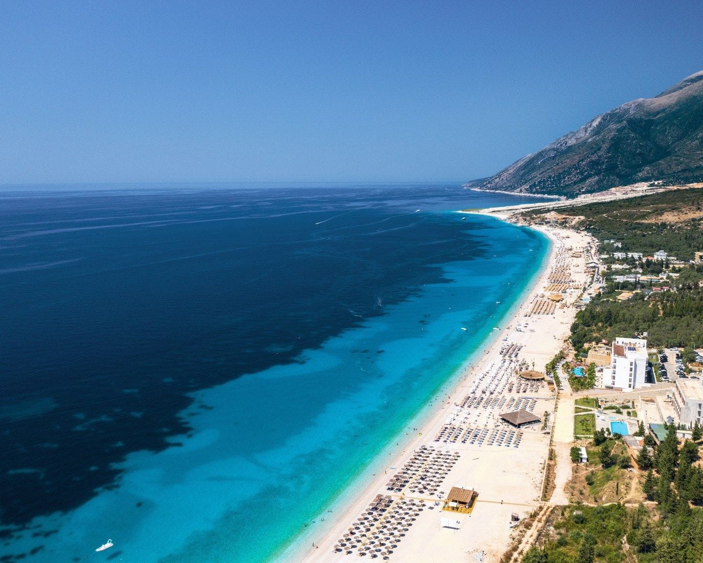 Drymades Beach near Dhërmi, Albania, with a long stretch of turquoise shoreline, sunbeds, resorts, and mountains in the background.