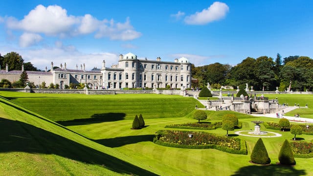 View of Powerscourt Estate in Ireland with its grand mansion, manicured gardens, green lawns, and blue sky with scattered clouds.