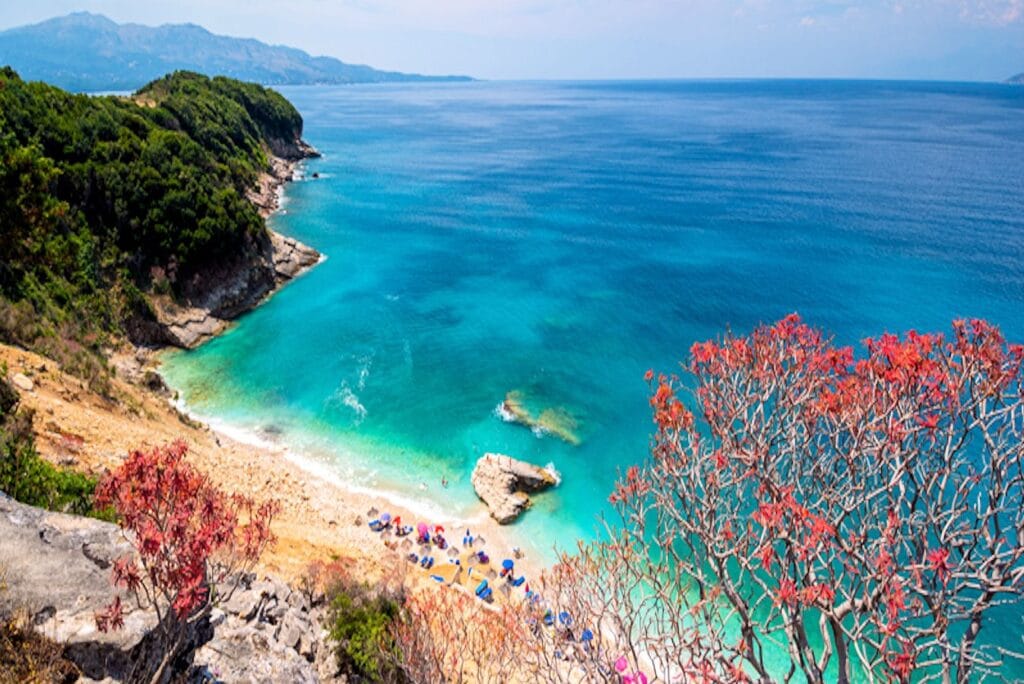 Aerial view of Pulebardha Beach in Albania with clear turquoise water, rocky shoreline, and rows of umbrellas on a pebbled beach.