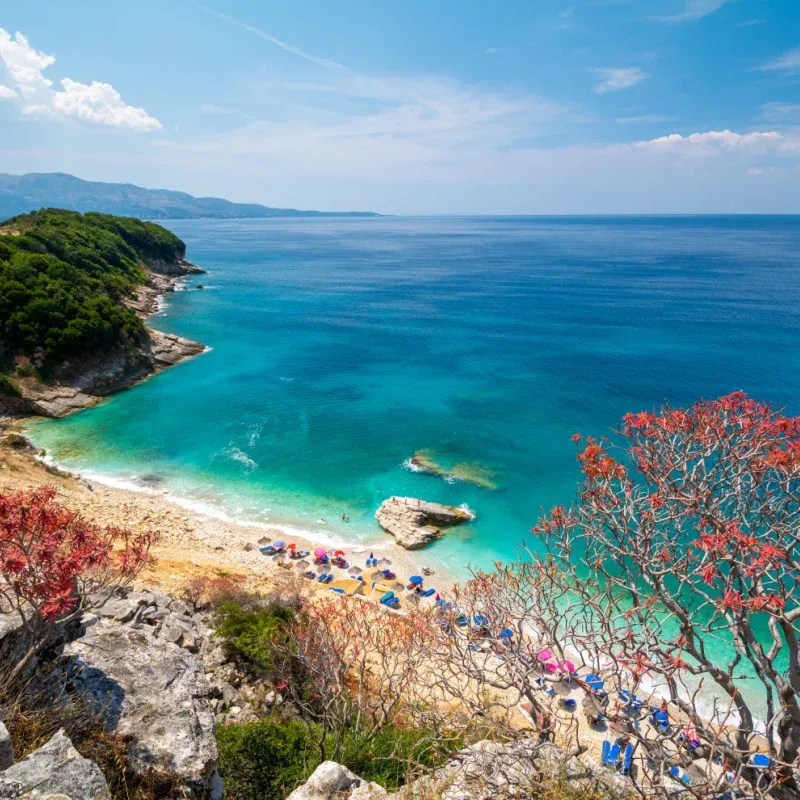 View of Pulebardha Beach near Ksamil, Albania, with clear turquoise water, white cliffs, beach umbrellas, and a rocky coastline.