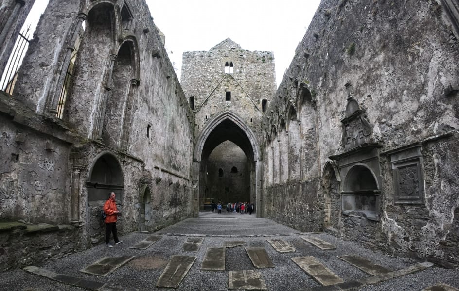 Interior of the medieval Rock of Cashel in Ireland, featuring weathered stone walls, arched windows, and tomb slabs on the gravel floor, with tourists exploring the historic ruins.