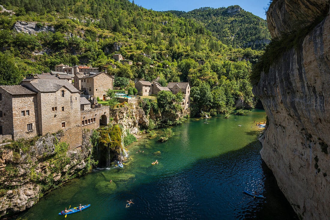 Riverside village of Saint-Chély-du-Tarn with stone houses, waterfalls, and people kayaking in clear water.