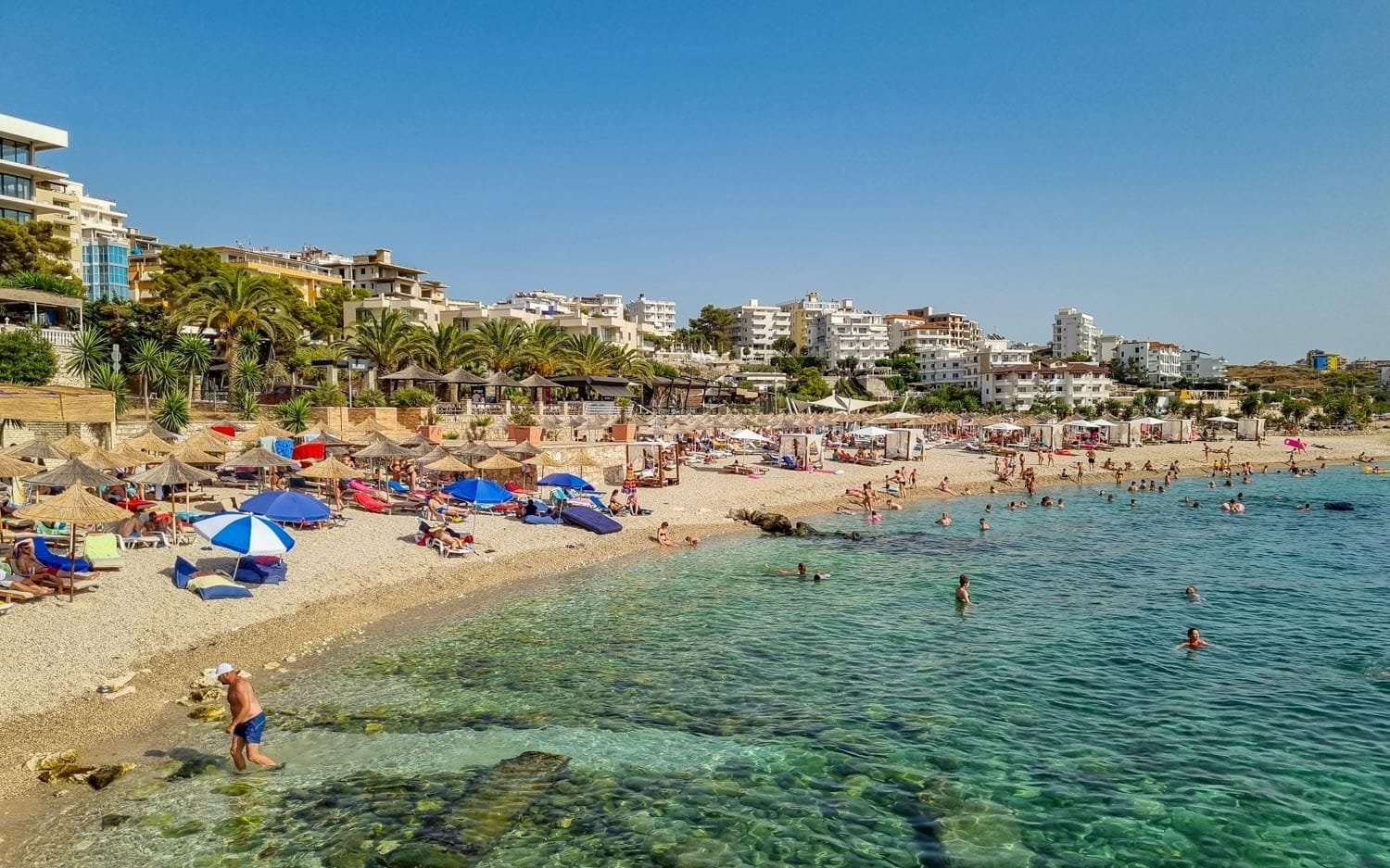 Busy day at Saranda City Beach in Albania with sunbeds, umbrellas, palm trees, and clear shallow water along the waterfront promenade