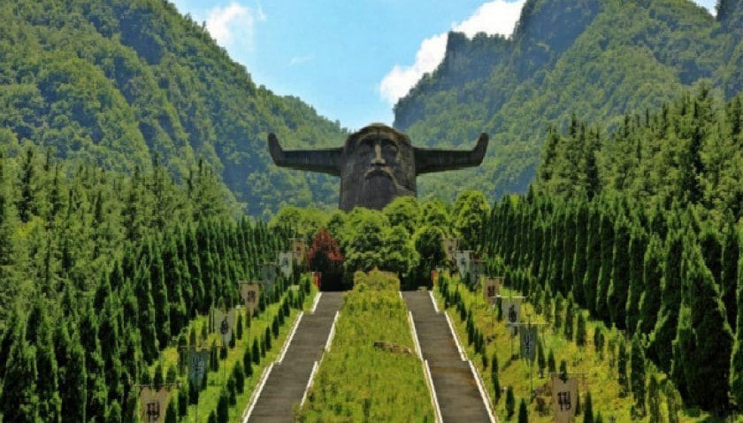 Monumental statue of the mythological figure Shennong with outstretched arms at the end of a tree-lined path in Shennongjia National Park, Hubei, China, set between densely forested mountain slopes.